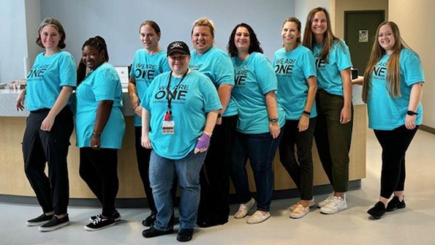 A large group of staff members lean against a front desk while wearing We Are One t-shirts