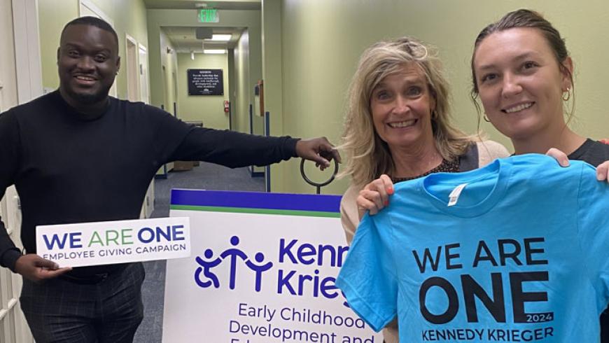 Three staff members smile while standing in a hallway holding We Are One signs and shirts.