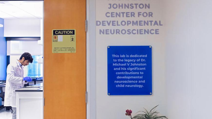 A male researcher works inside the Johnston Lab. The image is taken from a hallway, with one door to the lab open.