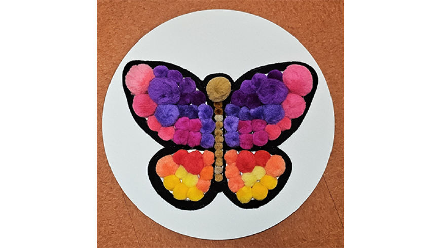 A butterfly design made from colorful pom‑poms in shades of pink, purple, yellow, and orange on a white circular background.