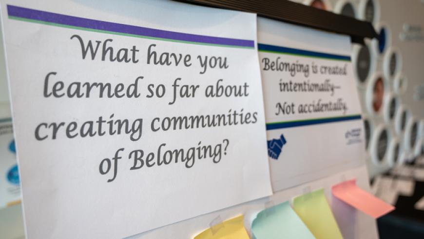 Two conference signs on a wall; the front asks, What have you learned about creating communities of Belonging?, and a second sign reads, Belonging is created intentionally—not accidentally.