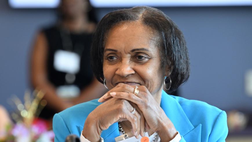 A woman with her hands clasped sits at a table and listens to a conference presentation.