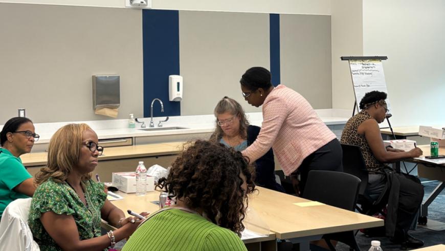 Participants seated around tables listen and engage in a workshop discussion in a classroom-style meeting room.