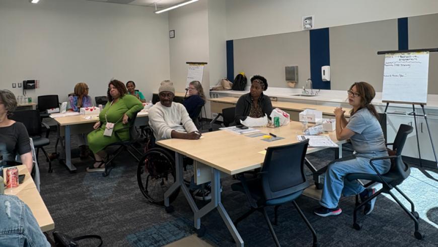 Workshop participants sit at tables in a meeting room while a facilitator leans over to assist an attendee during a group activity.