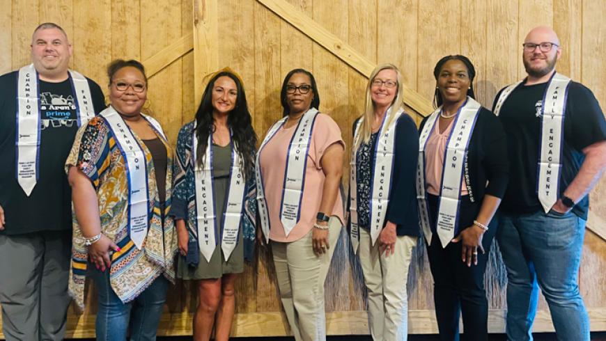 Six people wearing “Family Engagement” and “Champion” stoles stand side by side in front of a wood-paneled wall with barn-style trim