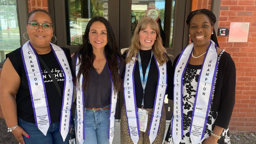 Four people wearing “Family Engagement” and “Champion” stoles stand together outside a brick building entrance, posing for a group photo.