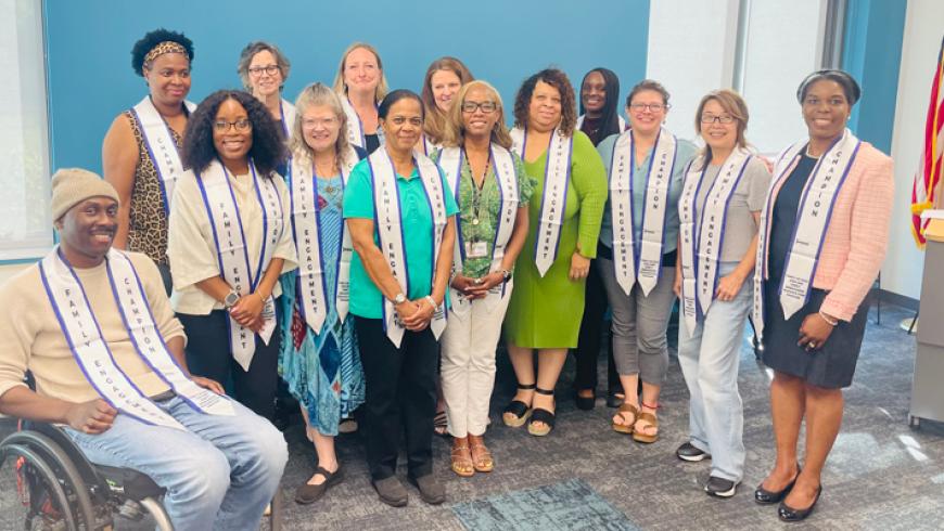 Group photo of 13 people standing and seated in a meeting room, many wearing white ceremonial stoles with printed text.