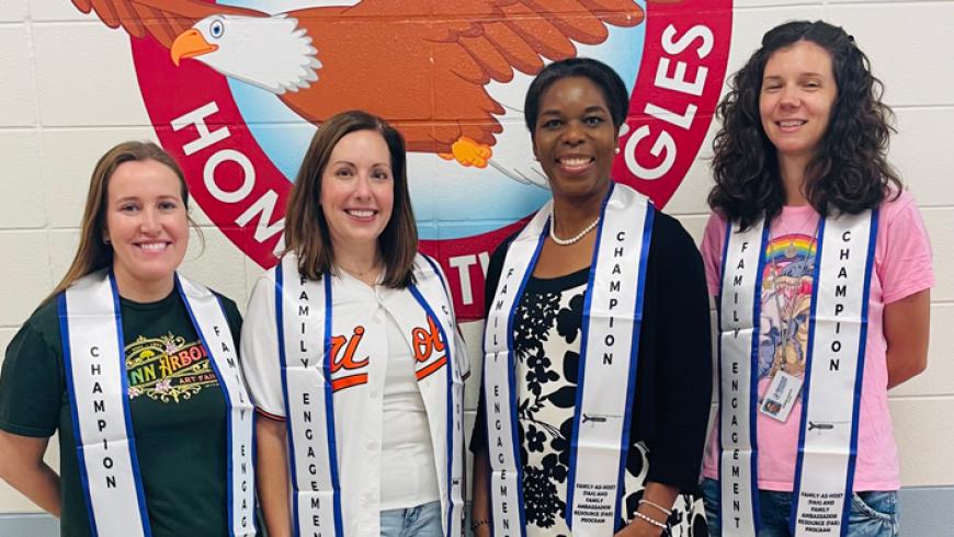 Four people wearing Family Engagement and Champion stoles stand in front of the Talbot Springs Elementary School wall mural, featuring an eagle mascot.