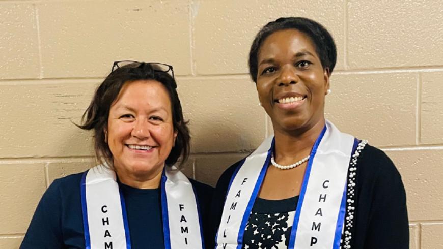 Two people wearing “Family Engagement” and “Champion” stoles stand together in front of a beige cinderblock wall, posing for a photo.