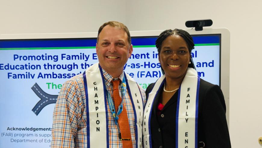 Two people wearing Family Engagement and Champion stoles stand in front of a presentation screen, posing for a photo.